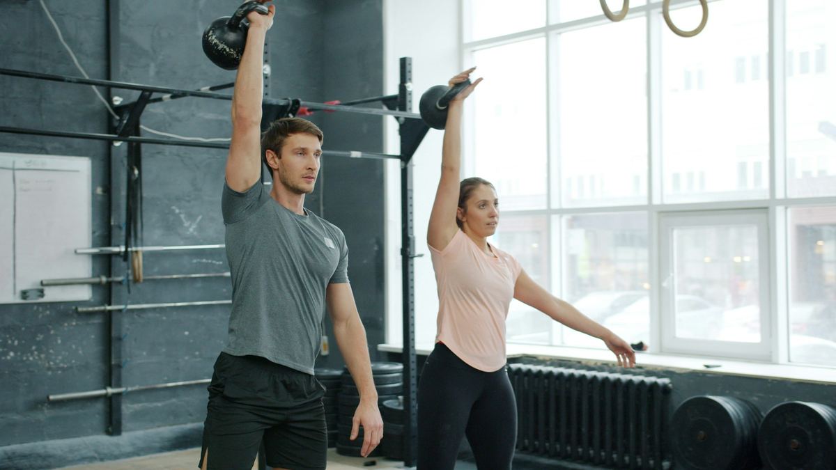 Een man en een vrouw houden elk een kettlebell boven het hoofd in een lichte gym.