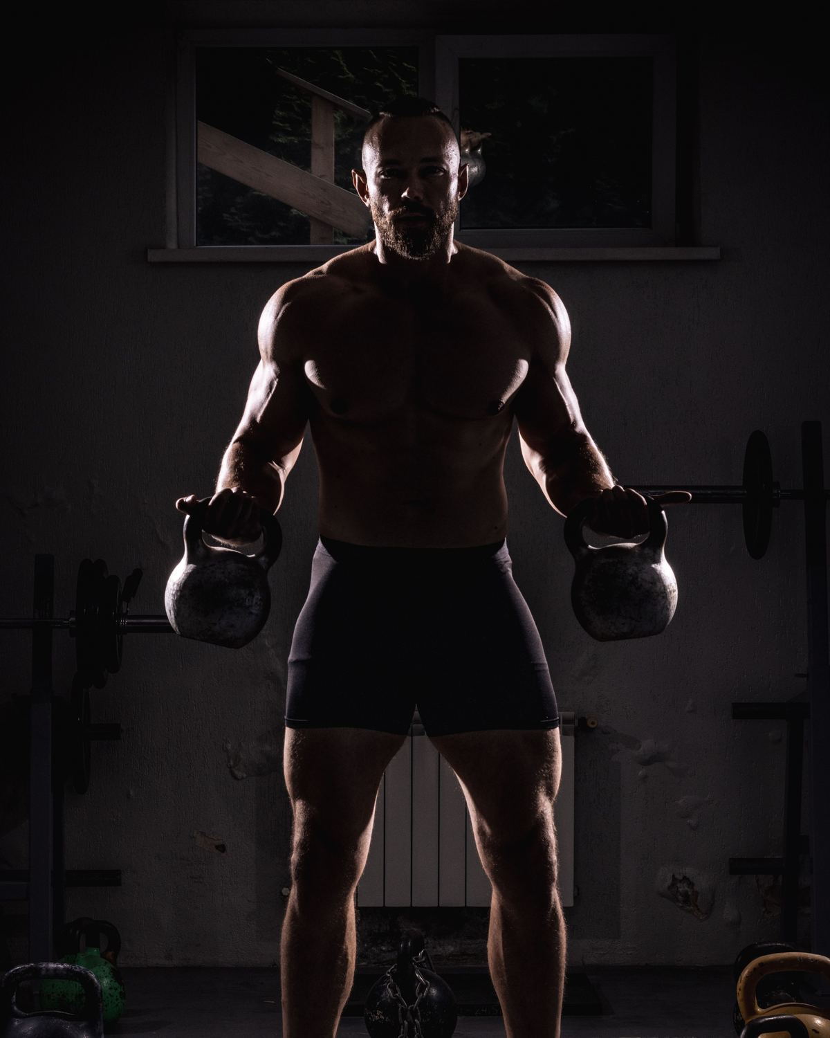 Een atleet staat in een donkere gym met in elke hand een kettlebell.